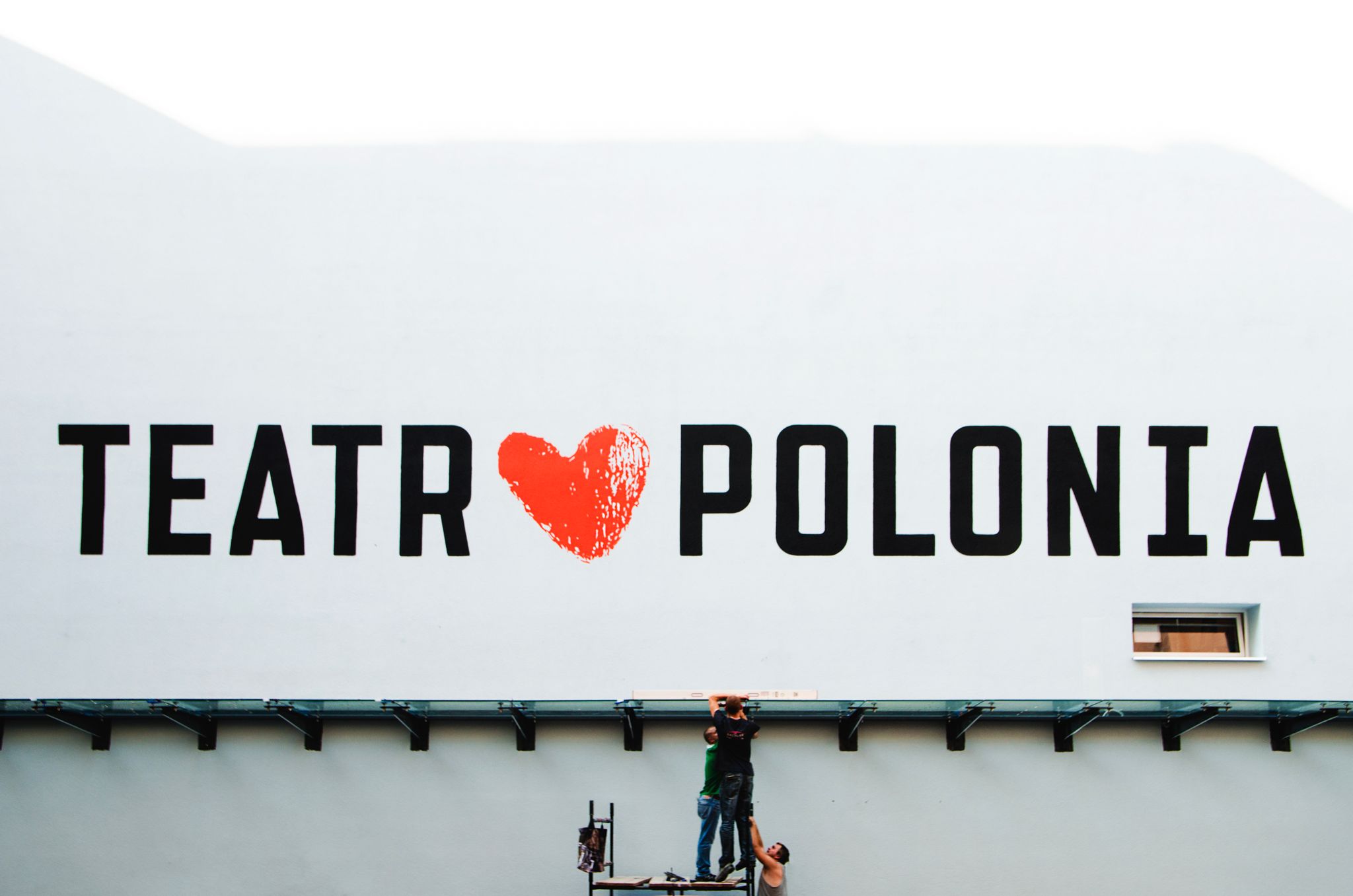 Workers on a lift painting the 'Teatr Polonia' sign on a large building exterior.